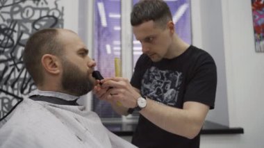 Berber berber dükkanında tıraş makinesiyle sakallı adam buduyor. Saç şekillendirme işlemi. Sakallı bir erkeğin sakalını kesen bir kuaförün yakın çekimi..