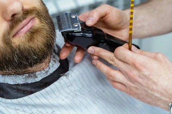 Barbier schneidet bärtigen Mann mit Rasierapparat im Friseursalon. Frisurenprozess. Nahaufnahme eines Friseurs, der einem bärtigen Mann den Bart schneidet. — Stockfoto