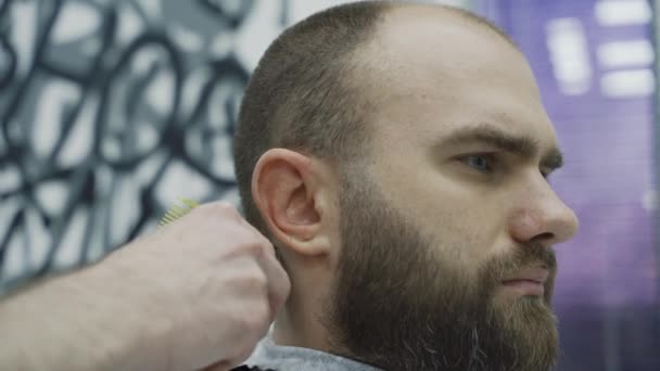 Barbeiro cortando homem barbudo com máquina de barbear na barbearia. Processo de penteado. Close-up de um cabeleireiro cortando a barba de um macho barbudo . — Vídeo de Stock