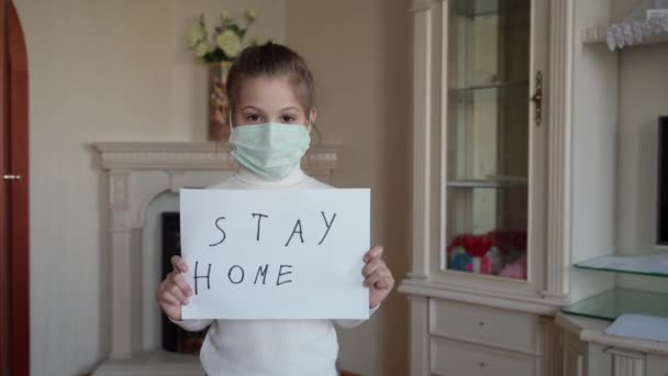Оставайтесь дома концепции. Маленькая девочка в маске держит табличку говоря остаться дома для защиты от вирусов и заботиться о своем здоровье от COVID-19 . — стоковое видео