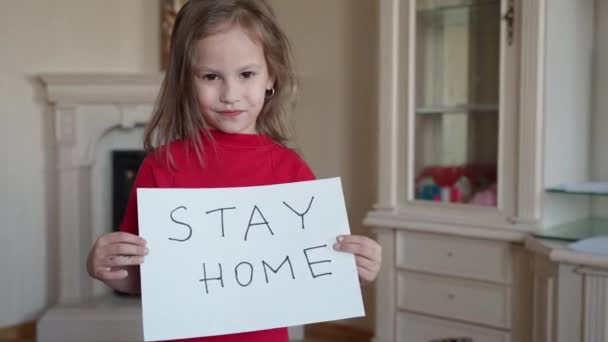 Bleiben Sie zu Hause. Kleines Mädchen in rotem N-Shirt, das ein Schild mit der Aufschrift "Bleiben Sie für den Virenschutz zu Hause und kümmern Sie sich um ihre Gesundheit von COVID-19" hochhält. — Stockvideo