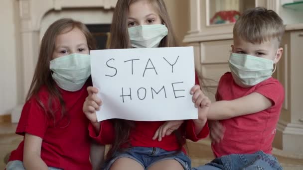 Familia estancia en el concepto de casa. Tres niños enmascarados sostienen letrero diciendo que se queden en casa para la protección contra el virus y cuiden su salud de COVID-19 . — Vídeo de stock