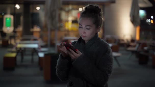Привлекательная молодая хипстерша в пальто, используя мобильный телефон, стоящий на улице ночного городка, счастливо реагирует на сообщение. молодая женщина пишет смс, разговаривает по смартфону на улице . — стоковое видео