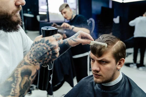 Attraktive Männer bekommen im Friseursalon einen modernen Haarschnitt. Tätowierter Barbier benetzt Haare mit Spray und kämmt sie. — Stockfoto