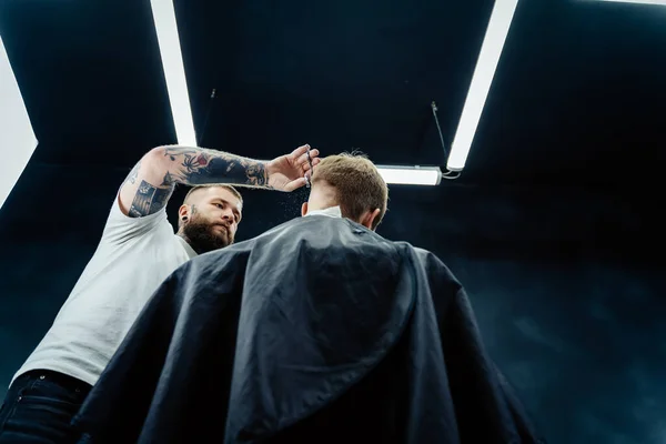 Tätowierter Barbier schneidet dem Kunden mit einer Schere die Haare. Aus nächster Nähe. Attraktive Männer bekommen im Friseursalon einen modernen Haarschnitt. Ansicht von unten. — Stockfoto