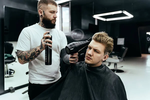 Barber makes hair styling with hair spray after haircut at the barber shop. Young handsome Caucasian man getting a haircut in a modern hairsalon.