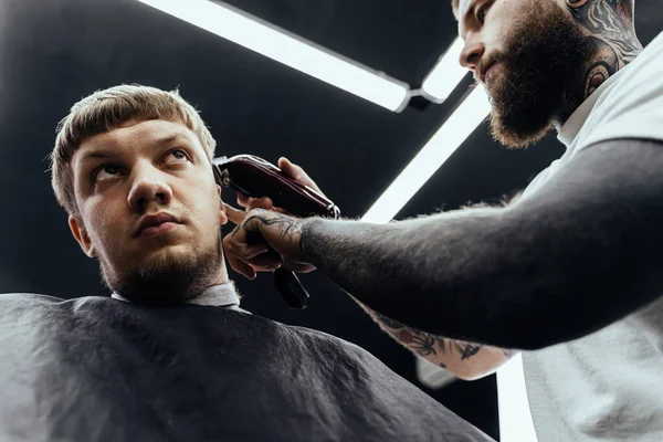 Männlicher Haarschnitt mit Rasiermesser. Tätowierte Friseurin schneidet Klienten im Friseursalon mit Haarschneidemaschine. Mann frisiert mit Elektrorasierer. Ansicht von unten. — Stockfoto