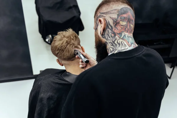 Manlig frisyr med elektrisk rakkniv. Tatuerad frisör gör frisyr för klienten i frisörsalongen med hjälp av hårklippare. Man frisör med elektrisk rakapparat. — Stockfoto