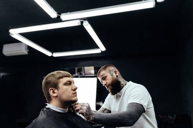 Berber dükkanında tıraş makinesiyle sakallı adam buduyor. Saç şekillendirme işlemi. Saç stilisti sakallı bir erkeğin sakalını kesiyor..