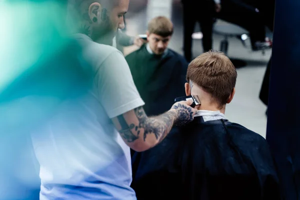 Male haircut with electric razor. Tattooed Barber makes haircut for client at the barber shop by using hairclipper. Man hairdressing with electric shaver.