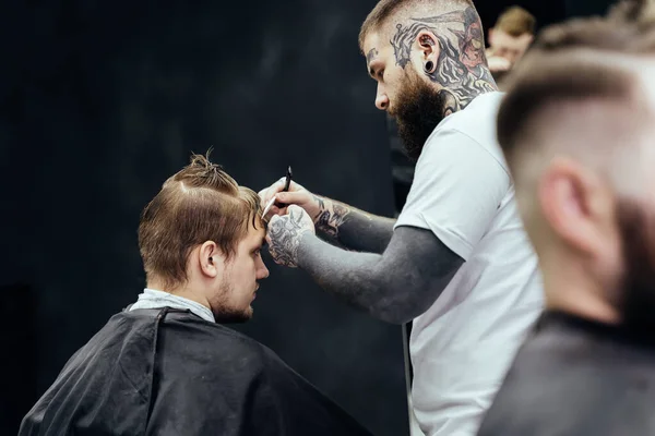 Professioneller tätowierter Friseur mit rasiermesserscharfen Haaren. Attraktive Männer bekommen im Friseursalon einen modernen Haarschnitt. Nahaufnahme. — Stockfoto