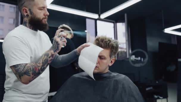 魅力的な男性は理容室の店で現代的な散髪を得ています。青いスプレーで顧客に髪を染めるバーバー. — ストック動画
