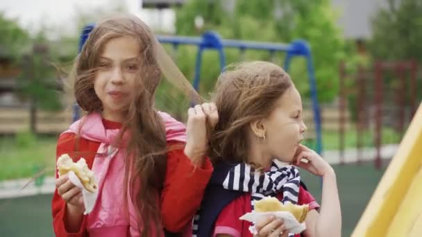 Duas meninas comendo sanduíches ao ar livre no parque infantil. Dia ventoso de verão. O bloqueio e a quarentena terminaram . — Vídeo de Stock