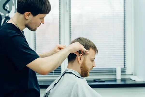 Tätowierter Barbier schneidet dem Kunden mit einer Schere die Haare. Aus nächster Nähe. Attraktiver Mann bekommt im Friseurladen einen modernen Haarschnitt. — Stockfoto