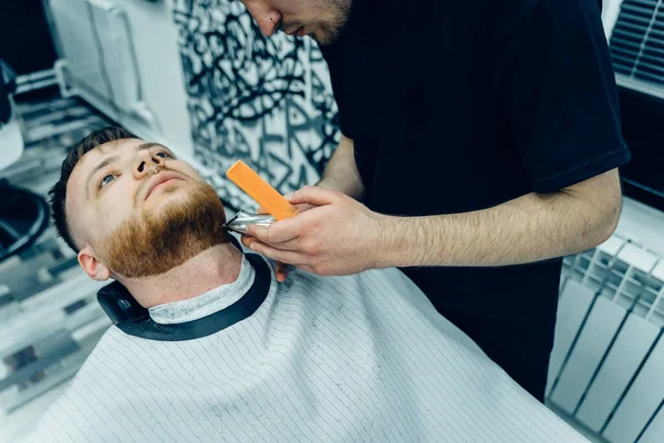 Barbier schneidet bärtigen Mann mit Rasierapparat im Friseursalon. Frisurenprozess. Nahaufnahme eines Friseurs, der einem bärtigen Mann den Bart schneidet. — Stockfoto