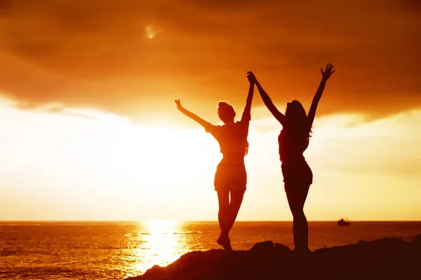 Две счастливые девушки с поднятыми руками на фоне заката моря — стоковое фото