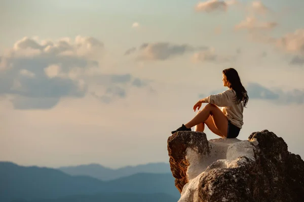 Девочка сидит на большой скале на фоне заката и гор — стоковое фото