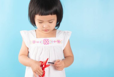 Elinin etrafındaki kaslardan bir şeyler kesmeyi öğrenmek için elinde kırmızı plastik makas tutan tatlı Asyalı kız. Mavi bir arkaplanda ve kopyalama alanı.