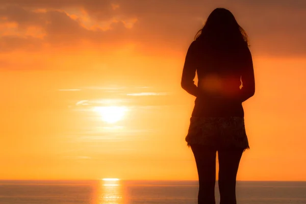 Young woman stands looking out to sea at the sunset