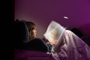 Geceleri rahat bir yatak odasında pijama giymiş kitap okuyan bir kız.