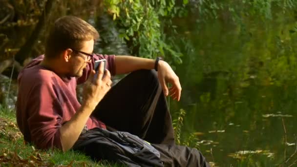 Muž psaní zpráv venku zavolá sledovat Video přehrávání virtuální hry ve slunečný den parku u turistickým batohem vody sedí na pozemní — Stock video