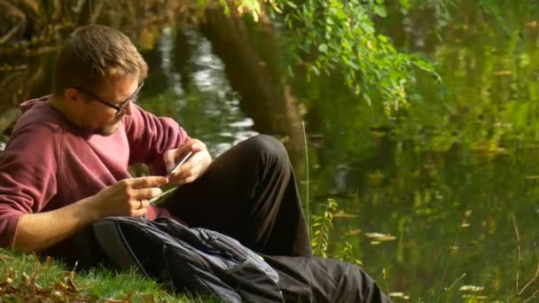 Mann tippt eine Nachricht ein und schaut auf den Bildschirm im Freien, während er im sonnigen Park am Wasser virtuelle Spiele anschaut — Stockvideo