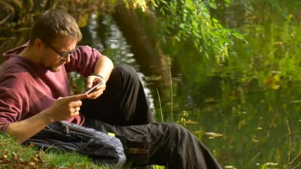 Mann tippt eine Nachricht auf den Bildschirm schaut Videospiele in sonnigen Tag Park durch das Wasser Tourist sitzt auf einem Boden ersetzt den Rucksack — Stockvideo