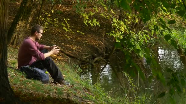 Ο άνθρωπος έρχεται κάνει μια κλήση πληκτρολογώντας ένα μήνυμα σε εξωτερικούς χώρους βλέποντας βίντεο παίζουν εικονικά παιχνίδια στο ηλιόλουστο πάρκο ημέρα από τα τουριστικά Backpacker νερό που κάθεται σε ένα έδαφος — Αρχείο Βίντεο