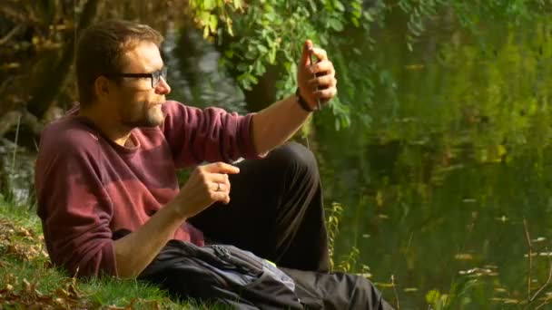 Man Makes a Video Call Waves His Hand Smiling Outdoors Sitting on a Ground in Sunny Day Park by the Water Backpacker Tourist Has been Leaned to His Elbow — Vídeos de Stock