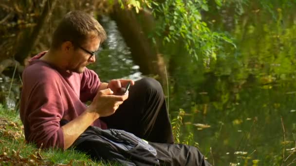 Un homme passe un appel Regarder une vidéo Parler à quelqu'un qui sourit dans un parc ensoleillé près de l'eau Randonneur touristique Happy Young Man is Sitting on a Ground — Video