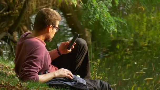 Man een bericht buiten te typen aan het meer kijken naar Video virtuele spelen in zonnige dag Park door de Water rugzaktoerist zittend op een grond — Stockvideo