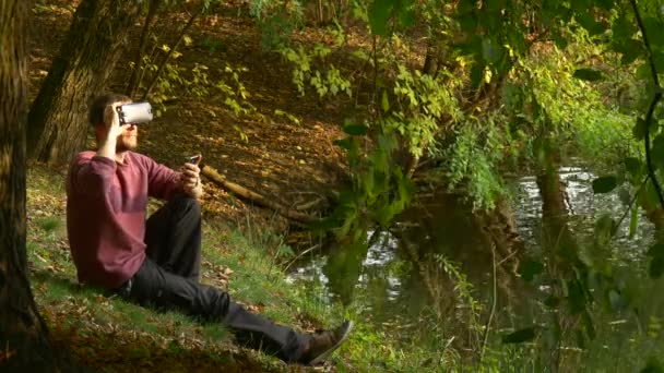 Man Puts on 360Vr Glasses Watching Video 360 Degrees Holding Mobile Phone Feels Real Turning His Head Playing Virtual Games in Sunny Day Park by Lake — Stock Video