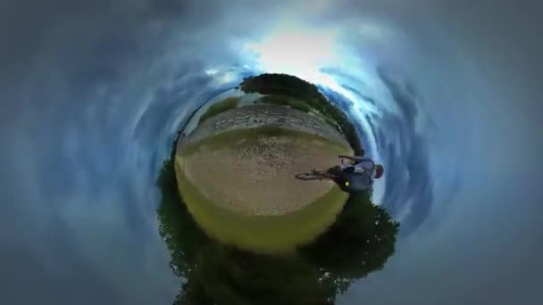 Kleiner winziger Planet 360-Grad-Tourist fährt Fahrrad entlang des Flusses Meer steinigen Ufer Backpacker reist warmen Herbst Tag bewölkten Himmel Tour nach Opole — Stockvideo