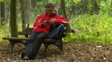 Adam gözlüklü bir mesaj Park sonbahar Tıklanma telefon ve gülümseyen izlerken Video oynarken sanal oyunlar oturan yapısı, kırık bir bankta yazmakta olduğunu