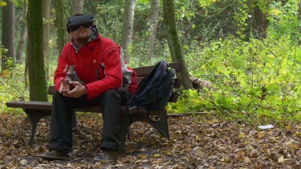 Człowiek w okularach 360vr jesienią Park, oglądanie wideo 360 stopni, gry wirtualne gry znaczki nogi rozglądając się czuje, gry prawdziwe siedząc na ławce — Wideo stockowe