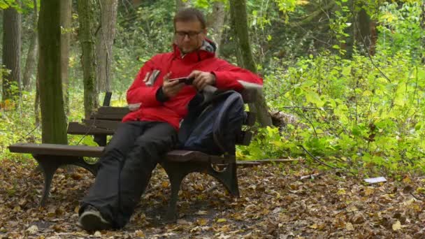 Hombre está sentado escribiendo un mensaje en el parque Otoño hace clic en el teléfono viendo video jugando juegos virtuales sentado en un banco roto en la naturaleza hojas caídas — Vídeo de stock