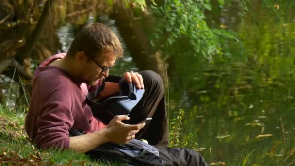 Young Man Holding 360Vr Glasses Clicks the Phone Park by the Water Tourist Looks Into the Glasses Watching Video 360 Degrees Playing Virtual Games Sunny — ストック動画