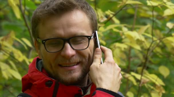 Mannen i Eyelasses är talande på Smartphone utomhus och leende i höst dag i parken eller skogen gul och grön lämnar träden är bakom the Tourist i rött — Stockvideo