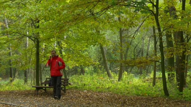 Турист разговаривает по мобильному телефону в парке Осень Завершает вызов и ищет что-то в рюкзаке сидя на скамейке у Природы Падшие листья — стоковое видео