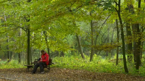 L'uomo si riposa sulla panchina a parlare sul cellulare nel parco Turismo autunnale seduto sulla panchina di legno abbandonata presso la natura in foglie cadute di giorno nuvoloso — Video Stock