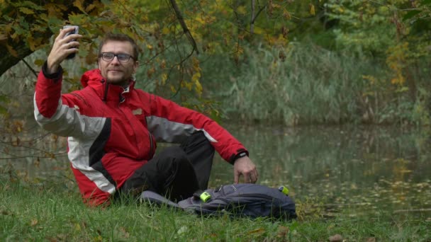 Турист делает селфи в парке в день водяного облака Молодой человек сидит на земле и улыбаясь отдыхает на берегу озера рюкзак размещен Она трава — стоковое видео