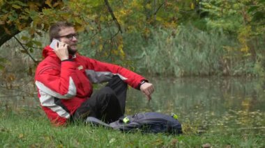 Turist üstünde hareket eden telefon Talking Park gülümseyen içinde bir yere su sonbahar bulutlu gün Backpacker gözlük geçirdiği zaman doğa tarafından üzerinde oturuyor