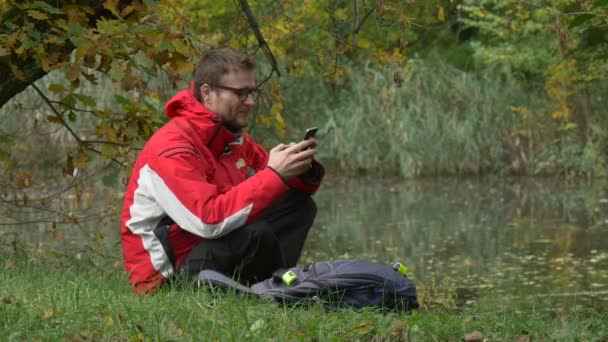 Tourist is Using Mobile Phone Texting Smiling Sitting in Park on a Ground by the Water in Autumn Cloudy Day Wild Ducks Are Swimming by Lake Surface — Stock Video