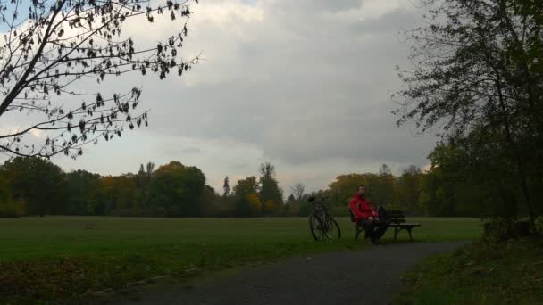 Radfahrer sitzt auf Bank am Fußweg Parkallee macht Rast in bewölkter Herbstdämmerung junger Mann in roter Jacke Fahrrad wird an der Holzbank zurückgelassen — Stockvideo