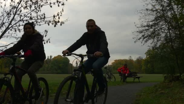 Люди в День семьи в парке Велосипедист сидит на скамейке Пара катается на велосипеде Силуэты и заклинает свой телефон, сидя на ногах — стоковое видео