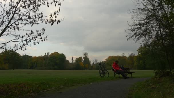 Ciclista entediado sentado no banco em Cloudy Day Park Man está descansando na natureza cliques telefone olhando ao redor paisagem outono bicicleta atrás do banco — Vídeo de Stock