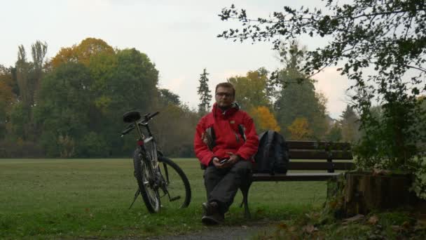 Znudzony człowiek klika telefonu SMS-y, oglądanie wideo 360 stopni wirtualne gry siedząc na ławce w parku w pochmurny dzień jesieni Backpacker w parku — Wideo stockowe