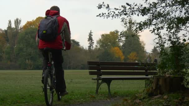 Turystyczna pochodzi z rowerów siedzi w dół na ławce w parku w pochmurne jesienne dzień rodziny jest Walking przez trawnik kobieta mężczyzna i dziecko Backpacker rowerzysta w parku — Wideo stockowe