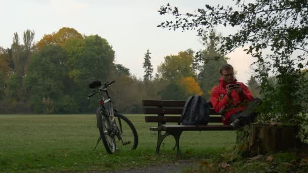 Man Makes a Call Puts on vr Headset Watching Video 360 Degrees Playing Virtual Games Sitting on the Bench in Park in Cloudy Autumn Day Backpacker in Park — ストック動画