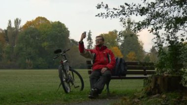 Adam bir görüntülü görüşme yapar ve gülümseyen konuşuyorsun bulutlu sonbahar gün Bisiklet parkta bankta sol arkasında tezgah bisikletçi Backpacker Park oturuyor